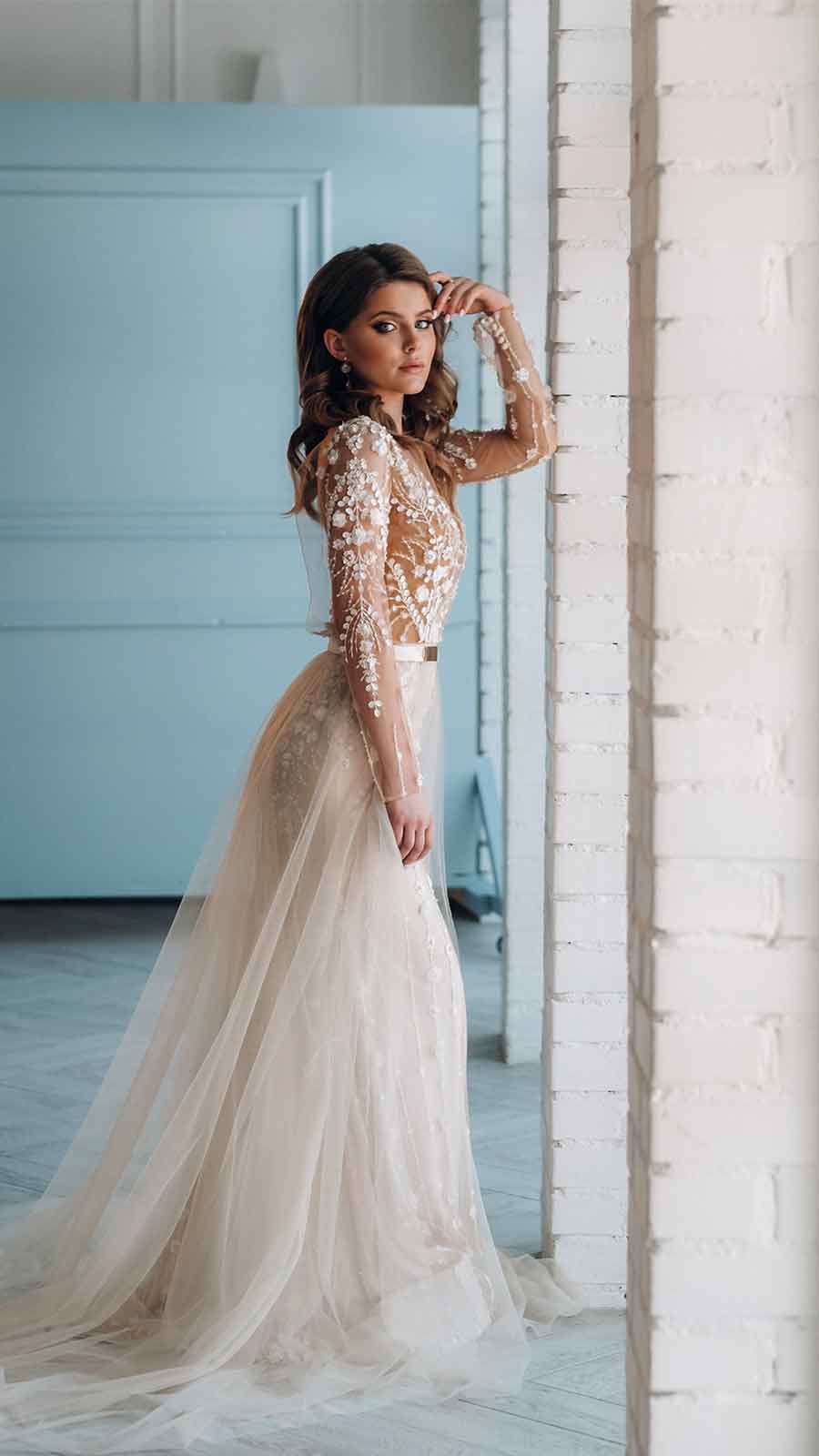 A woman in a long - sleeved wedding dress posing against a wall.