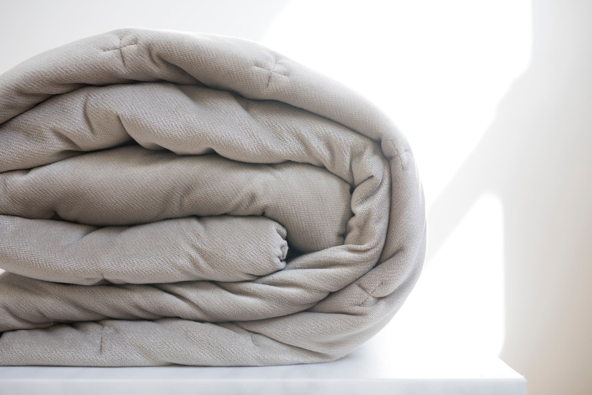 A rolled-up, light gray quilt or blanket is placed on a white surface with bright sunlight in the background.