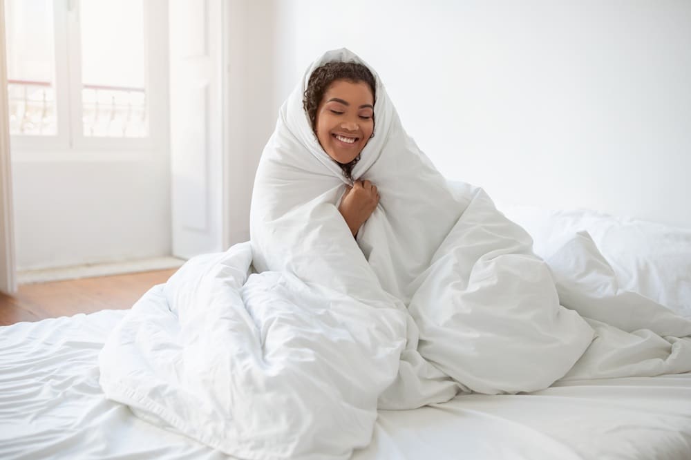A person sits on a bed wrapped in a white comforter, smiling, in a bright room with white walls and windows.