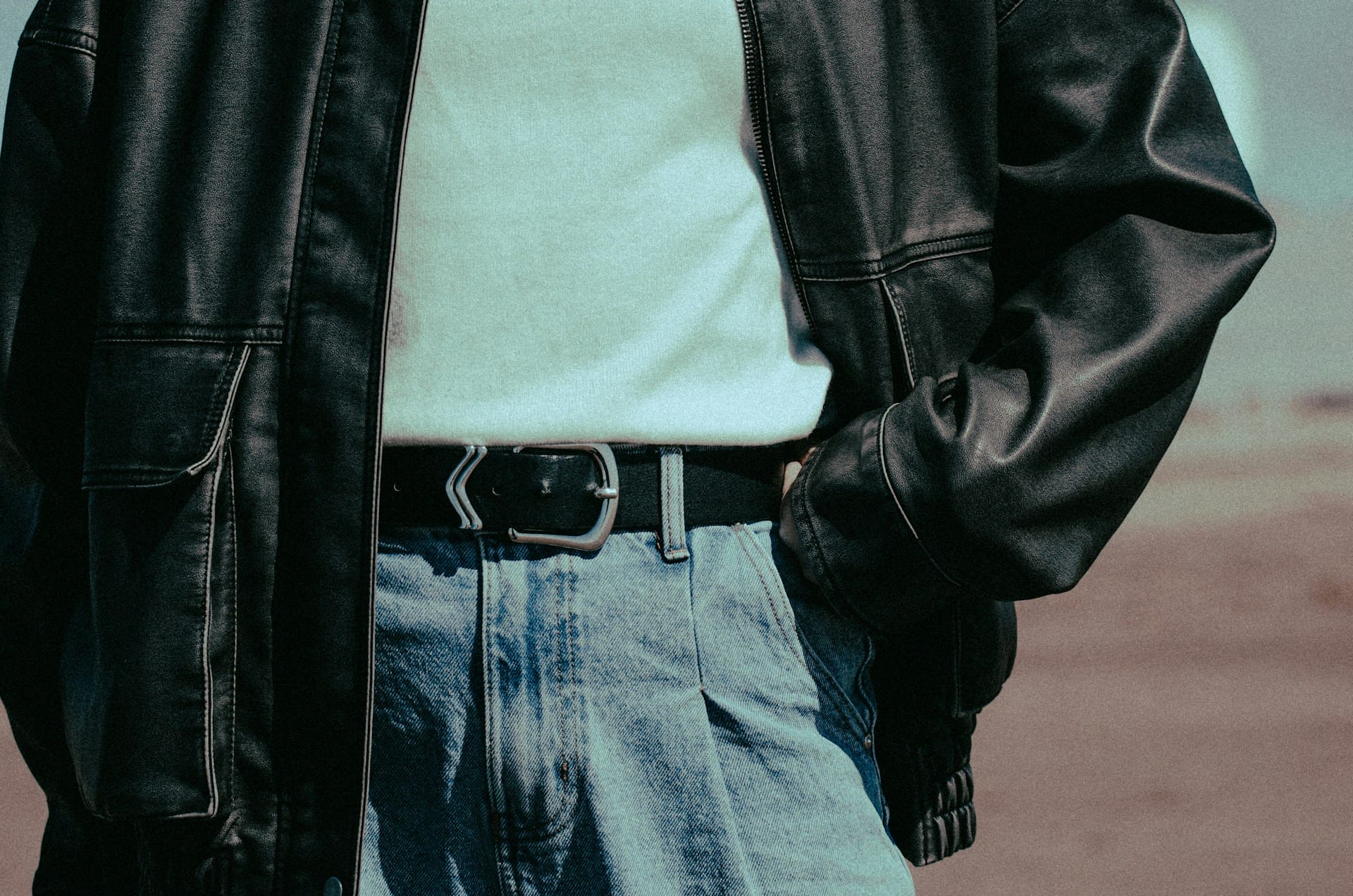 A person wearing a black leather jacket, white shirt, and blue jeans with a black belt, standing outdoors with one hand in their pocket.