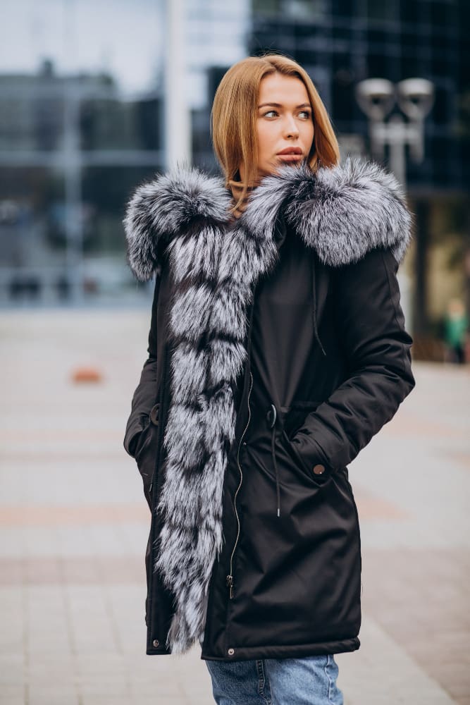 Woman in a black coat with a large faux fur collar and scarf standing outdoors in an urban setting, looking to the side.