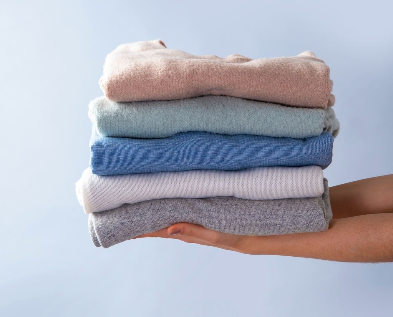 A person holds a neatly stacked pile of folded clothes in various pastel colors against a light blue background.
