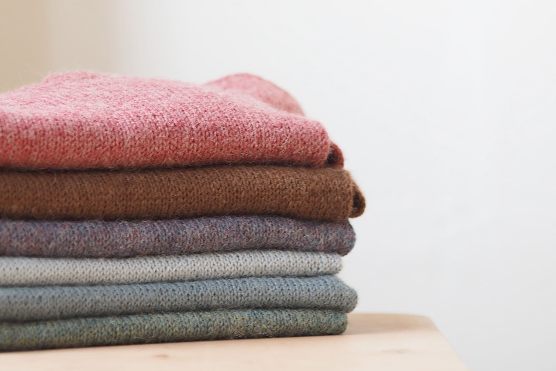 A neatly folded stack of wool sweaters in various colors, including pink, brown, gray, and blue, placed on a wooden surface against a plain background.
