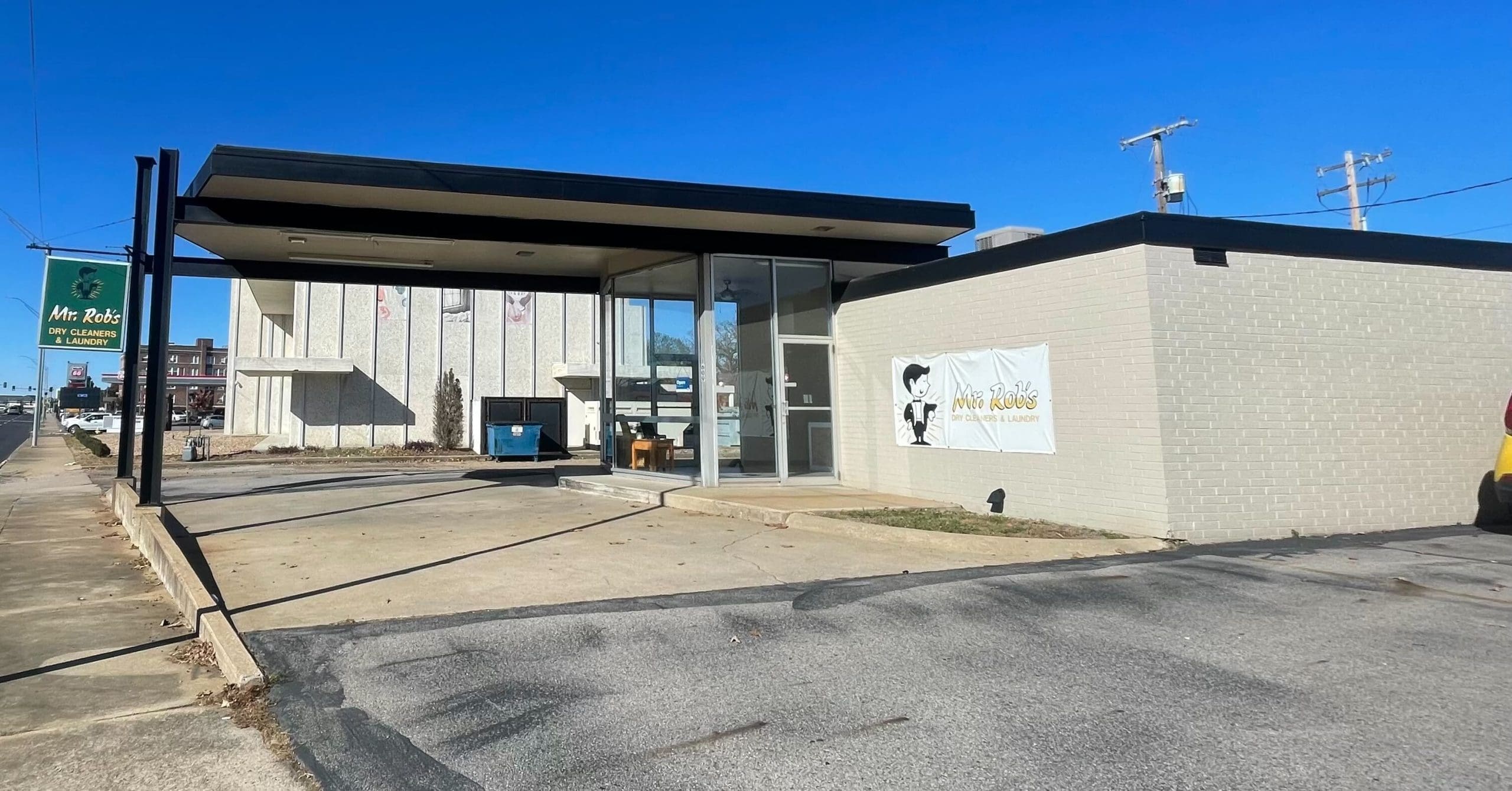 A small, modern building with glass walls and a flat roof houses “Mr. Rob’s Cleaners,” as indicated by signs on the exterior under a clear blue sky.