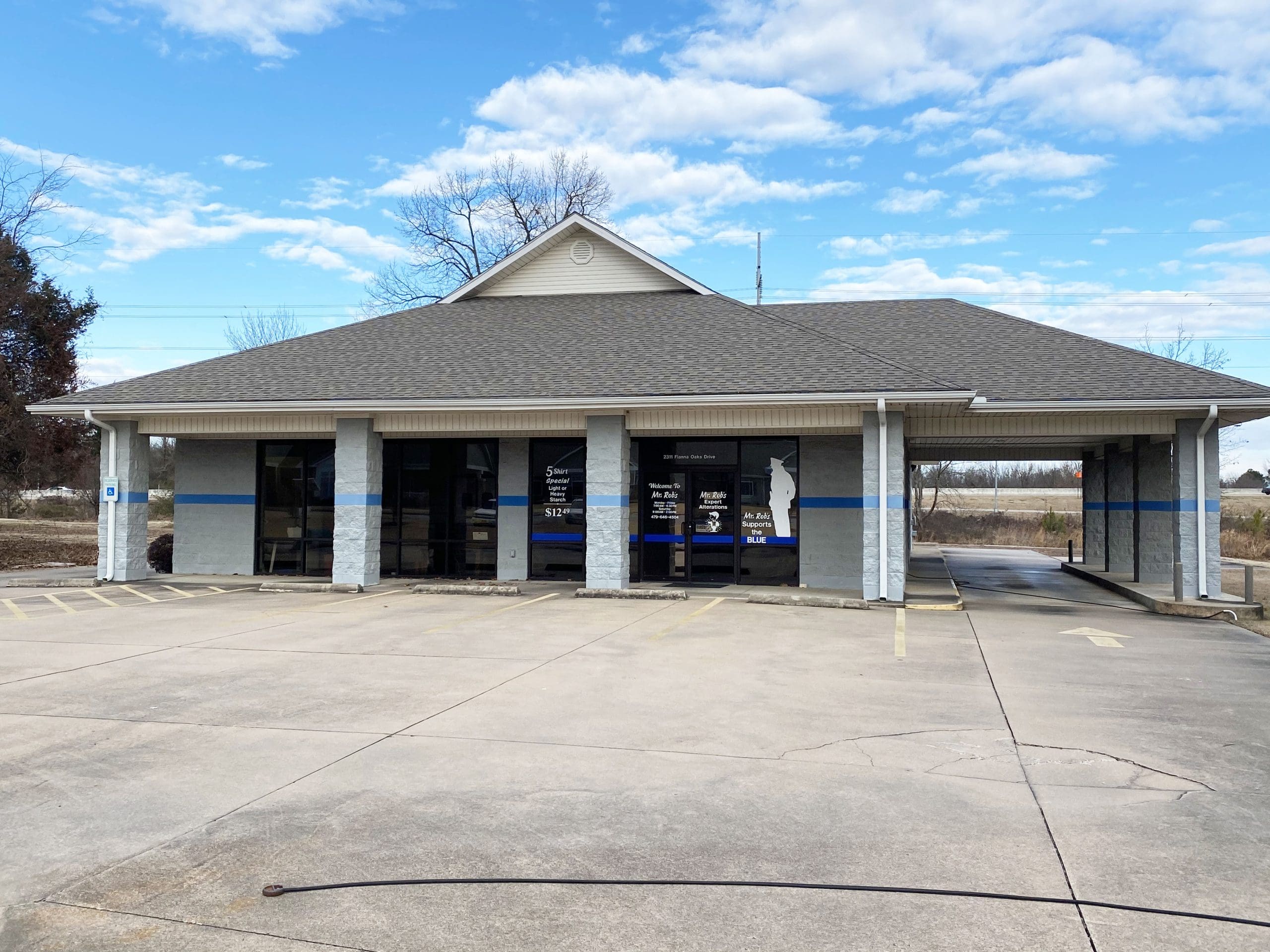 A one-story commercial building with a gray roof, blue and gray exterior, large windows, glass doors, and an empty parking lot in front under a partly cloudy sky.