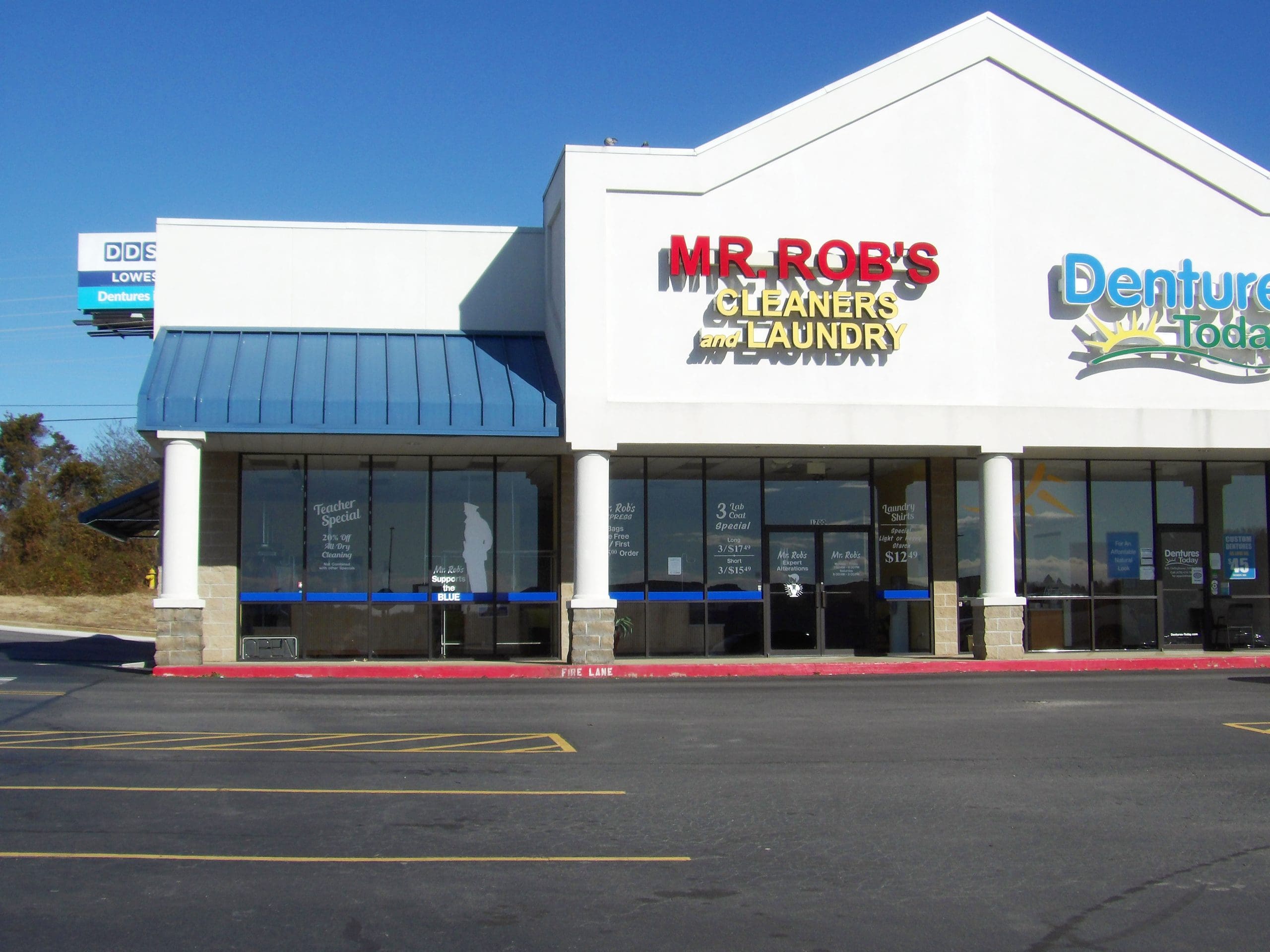 A strip mall storefront with signs for "MR. ROB'S CLEANERS and LAUNDRY" and "Dentures Today" on a clear, sunny day.