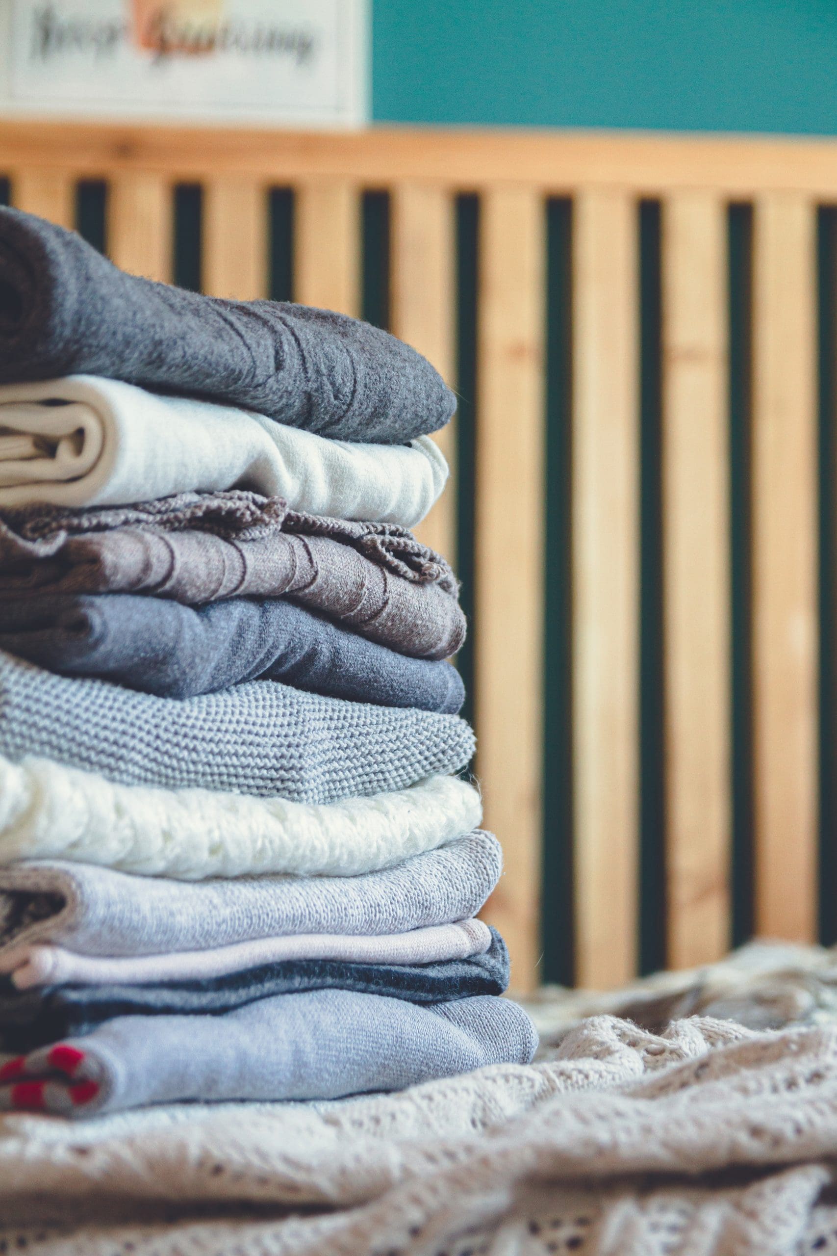 A neatly folded stack of assorted sweaters and knitwear is placed on a bed, with a wooden headboard in the background.