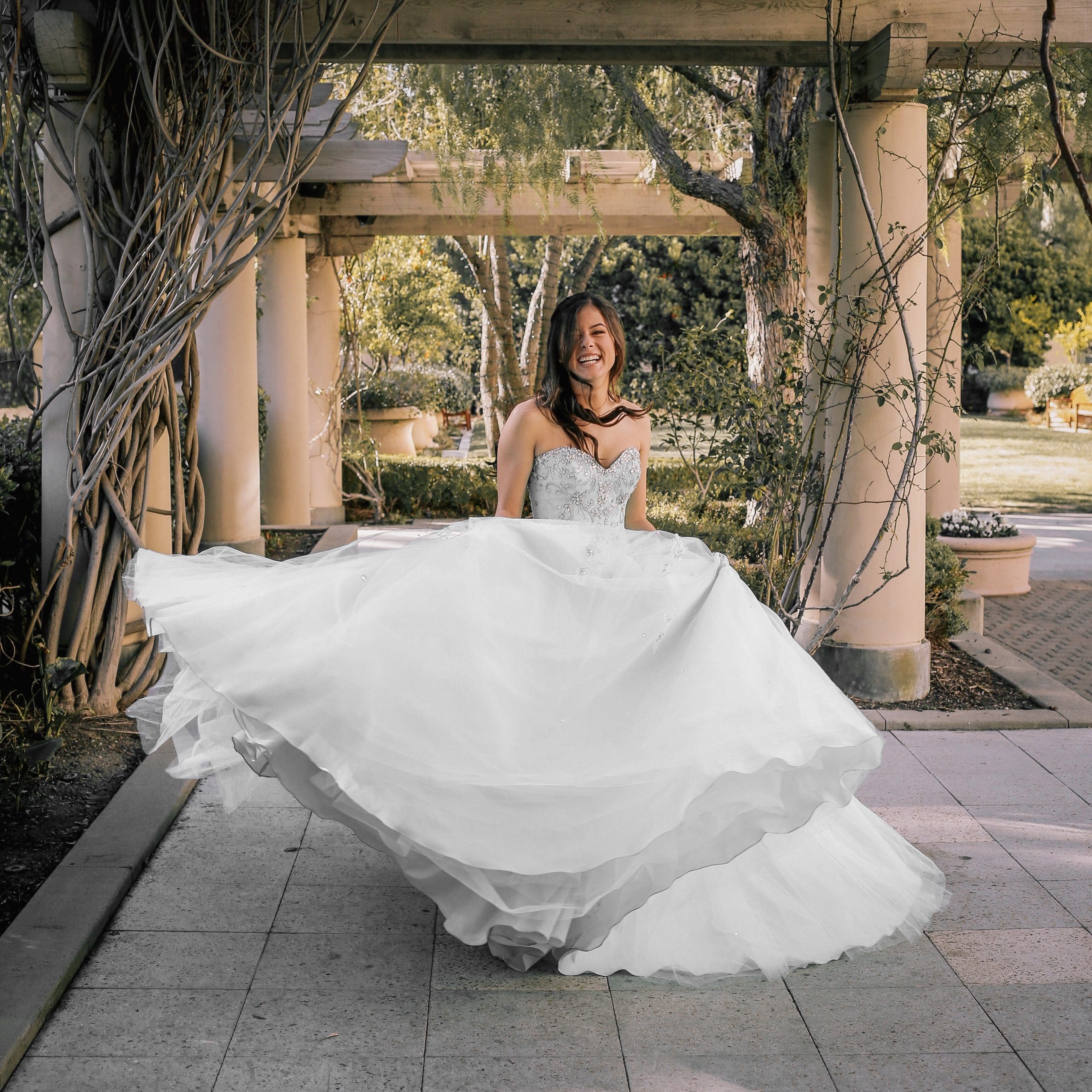 A woman in a white wedding dress twirls outdoors under a pergola, surrounded by greenery and sunlight.