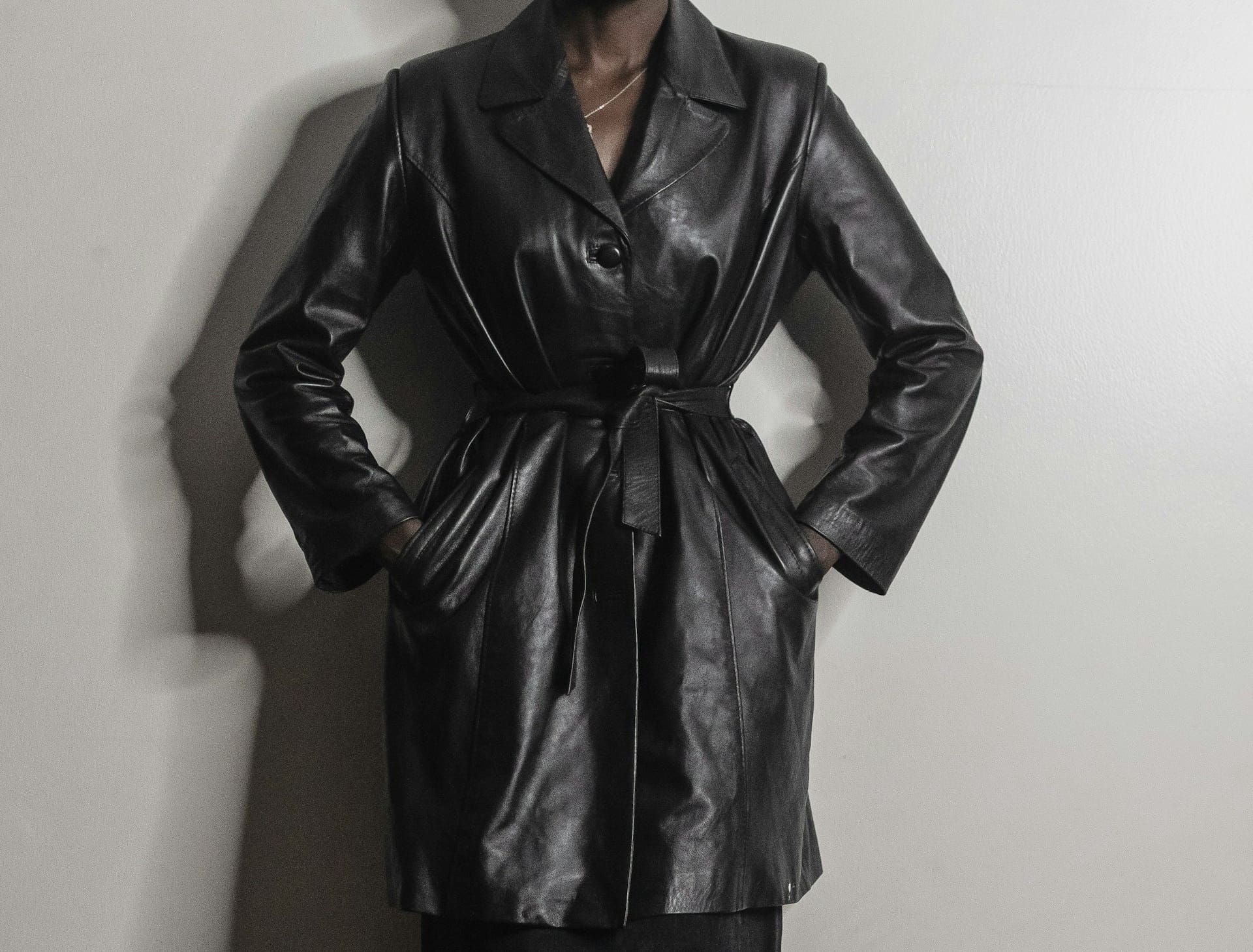 Person wearing a belted black leather trench coat with hands in pockets, standing against a plain light-colored wall.