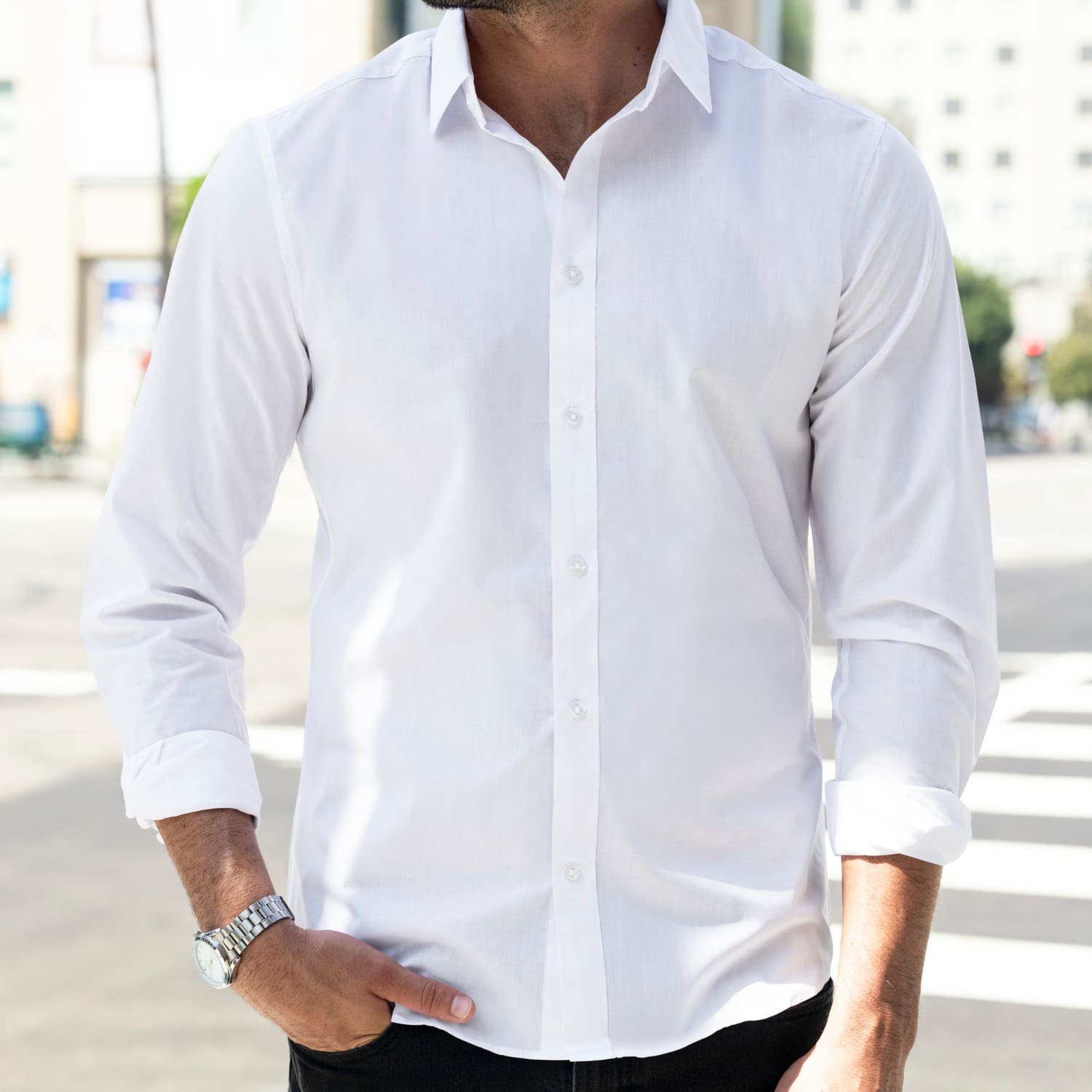 A man standing outdoors wearing a white button-up shirt with rolled-up sleeves, black pants, and a watch, with one hand in his pocket.